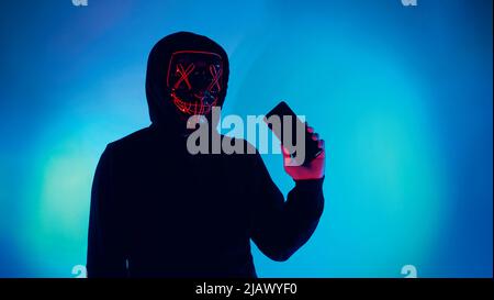 Anonymous hacker and face mask with smartphone in hand. Man in black hood shirt trying to hack personal data from mobile phone. Represent cyber crime Stock Photo