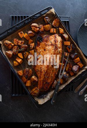 Oven dish with roasted and caramelized half salmon fillet. Cooked with potatoes and served on a baking tray on dark table background. Overhead view Stock Photo