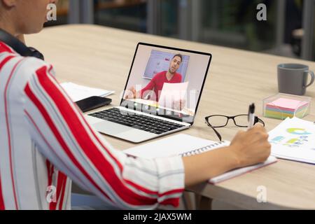 Caucasian businesswoman making notes while discussing with caucasian male colleague on video call Stock Photo