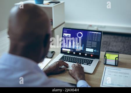 Rear view of african american businessman working on graph over laptop at desk in office Stock Photo