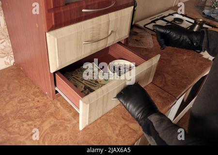 Robber in black outfit and gloves see in opened shelf in kitchen. The thief takes out the golden necklace from a kitchen shelf Stock Photo