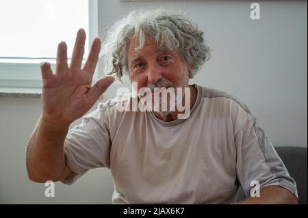Authentic senior man, grandpa, with grey curly hair waving with his hand towards the camera. Stock Photo