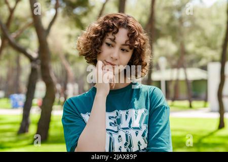 Redhead serious woman wearing green t-shirt standing on city park, outdoors. Touching chin thoughtful, thinking making important choice. Looking to gr Stock Photo