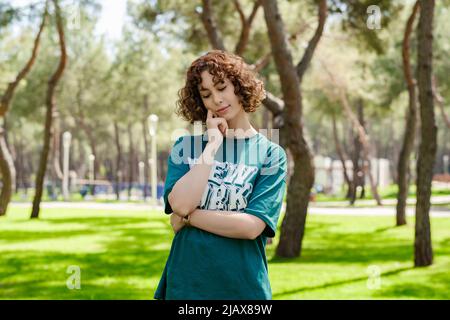 Young redhead woman wearing green t-shirt standing on city park, outdoors touching chin thoughtful, thinking making important choice. Stock Photo