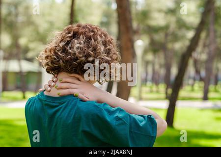Back view of young redhead woman wearing green tee standing on city park, outdoor feeling hurt joint shoulder back pain ache, fibromyalgia concept, cl Stock Photo