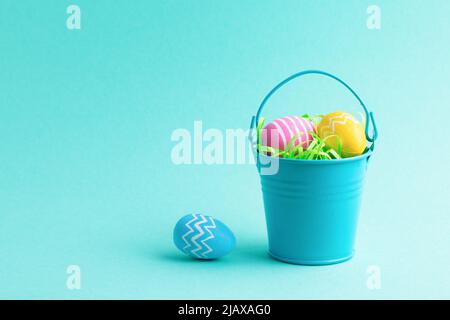 Spring holiday composition with colourful Easter eggs in small decorative bucket isolated on blue background. Front view, place for text. Stock Photo