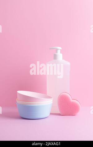 Dish washing composition with clean plates, heart sponge and cleaning detergent in plastic dispenser on pink background. Family care concept. Stock Photo