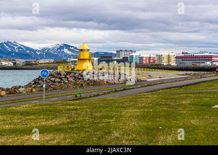 The yellow-painted Höfði lighthouse stands on a stone breakwater on the Sculpture and Shore Walk in Reykjavik, Iceland. An electric scooter leans against the railing, with the harbour and multicoloured apartment buildings visible against snow-capped mountains in the background. Stock Photo