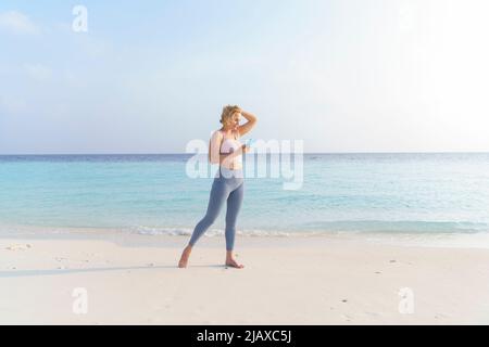 A sporty woman on the beach wearing headphones uses a smartphone. Stock Photo