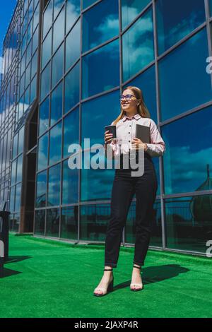 Beautiful woman manager in glasses and trendy clothes smiling on the office terrace, the girl drinks coffee and makes notes in a notebook. Stock Photo