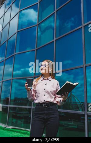 Beautiful woman manager in glasses and trendy clothes smiling on the office terrace, the girl drinks coffee and makes notes in a notebook. Stock Photo