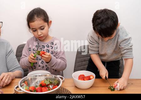 Cute children making fruit salad with mother in the kitchen. Kids cutting strawberries. Girl and boy learning to prepare meal from grandmother. Stock Photo