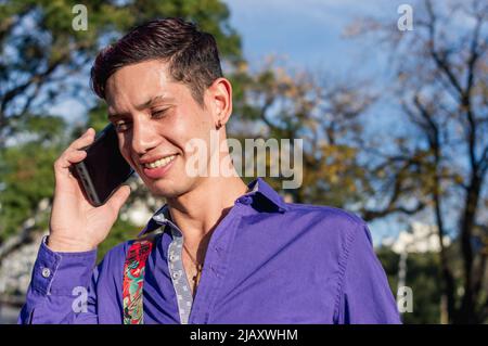 caucasian young latin man outdoors on a sunny day, wearing purple casual clothes, smiling and talking on the phone, technology concept, copy space. Stock Photo