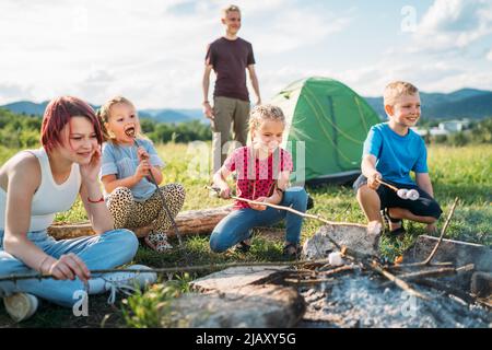 Boys and girls friends kids cheerfully laughing and roasting marshmallows on sticks over campfire flame near the green tent. Outdoor active time spend Stock Photo