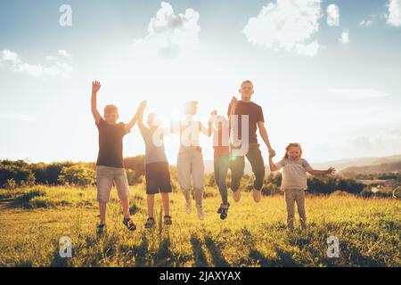 Six kids brothers and sisters teenagers and little kids funny jumping holding hands in hands on the green grass meadow with an evening sunset backgrou Stock Photo