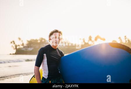 Portrait of a teen boy with Dental braces with surfboard goes for surfing. He is smiling and walking into the water. Happy childhood and active vacati Stock Photo