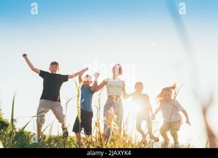 Selective focus brothers and sisters teenagers and little kids jumping holding hands in hands on the green grass meadow with an evening sunset backgro Stock Photo