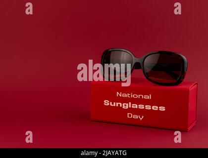 Brown sunglasses lie on a red background. Festive concept for national sunglass day. White text on the front of the box. Stock Photo
