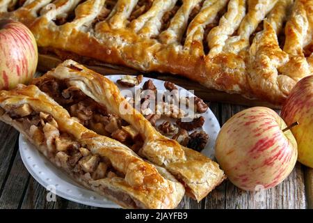 A piece of apple pie with walnuts strudel lies on a dessert plate against the background of a whole pie surrounded by chopped nuts and apples Stock Photo