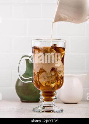 Cream being poured into an iced coffee in a tall glass mug Stock Photo