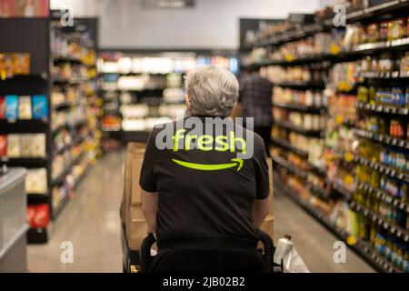 An elderly worker in an Amazon Fresh T-shirt pushes a cart loaded with pickup paper bags down the aisles inside an Amazon Fresh grocery store in ... Stock Photo