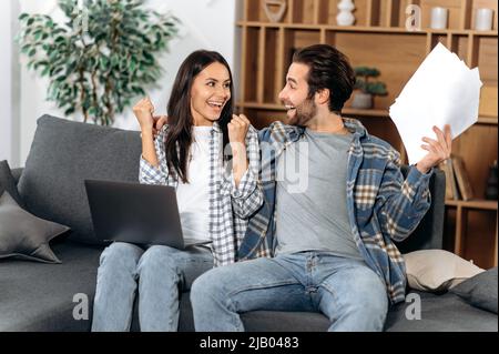 Happy family couple, in stylish clothes, sit on a sofa in the living room, use a laptop and documents, rejoice in big profits or buying real estate, gesticulate with hands, look at each other, smiling Stock Photo