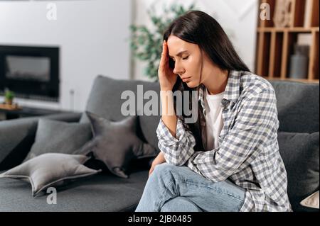 Headache, illness, personal experiences. Sad caucasian brunette woman, in casual clothes, sitting at home in the living room on the sofa, closed her eyes, experiencing stress, personal problems Stock Photo