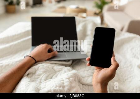 POV Of Man Using Smartphone And Laptop In Bedroom, Cropped Stock Photo