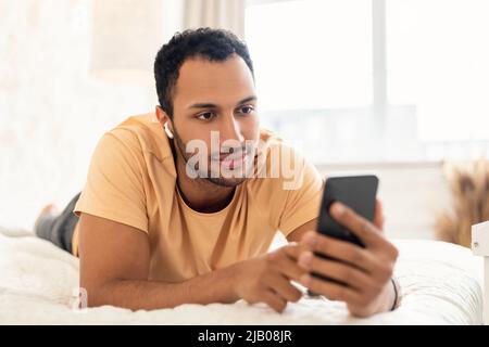 Middle Eastern Man Wearing Earbuds Using Mobile Phone In Bedroom Stock Photo