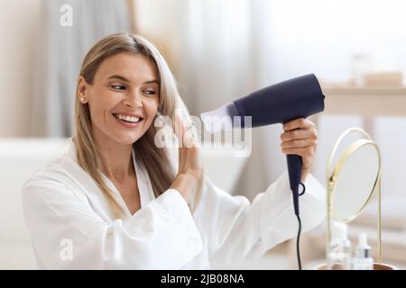 Attractive blonde woman using hair dryer at home Stock Photo