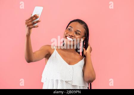Charming young black woman taking selfie with smartphone, filming content for her video blog on pink studio background Stock Photo