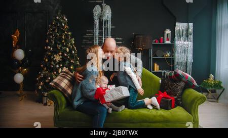 Christmas concept - family sitting on the sofa - dad giving others a gift - his wife and daughter kissing him in cheeks Stock Photo