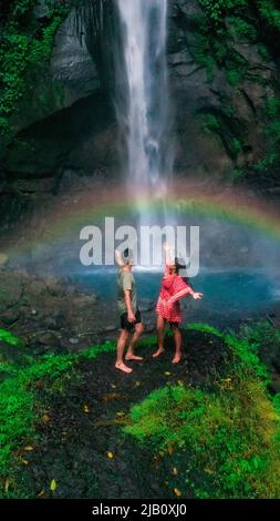 young travel couple standing on top a rock overlooking a dramatic waterfall with a natural real rainbow over the lagoon Stock Photo