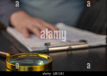 DEFOCUSED SHOT of a hand resting on a clipboard. A gold-rimmed magnifying glass is in the foreground. Adult male laying his hand on documents with han Stock Photo