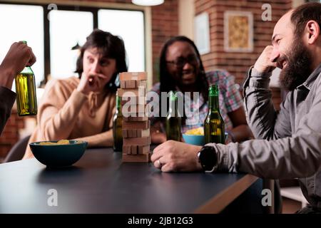Multiethnic happy friends sitting at table in living room while playing society games together. Diverse people having fun by enjoying snacks, beer and fun leisure time activities at home. Stock Photo
