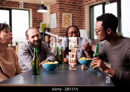 Multiethnic group of friends sitting at table in living room while playing together society games. Diverse people sitting at home while enjoying party game with wooden blocks and tower structure. Stock Photo