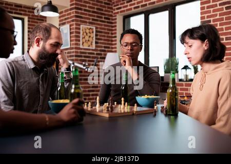 Pensive african american man thinking about next chess move while sitting at table. Focused confident person sitting at home in living room with multiethnic friends while playing strategy boardgames. Stock Photo