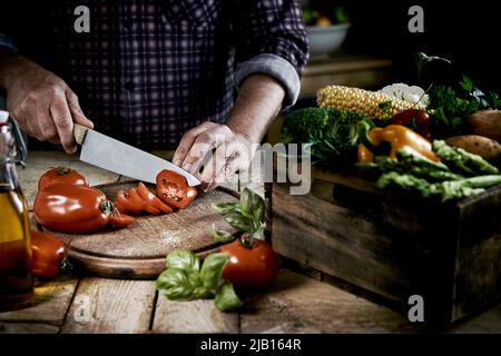 From above of crop anonymous male chef in checkered shirt slicing fresh ripe tomatoes on round shaped wooden cutting board at table with box of assort Stock Photo