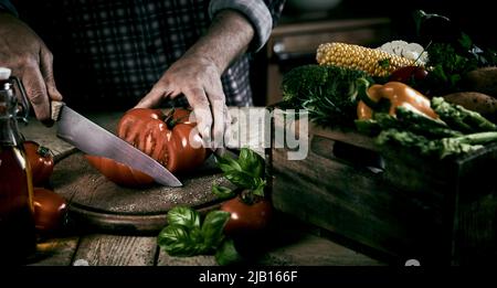 High angle of crop unrecognizable male chef cutting fresh ripe tomato on chopping board while standing at table with wooden box with assorted vegetabl Stock Photo