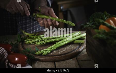 High angle of crop unrecognizable male chef peeling fresh ripe asparagus standing at wooden table with assorted vegetables in kitchen Stock Photo