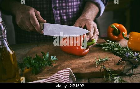 Crop unrecognizable male chef in checkered shirt cutting fresh bell peppers on wooden chopping board while standing at table with olive oil bottle and Stock Photo