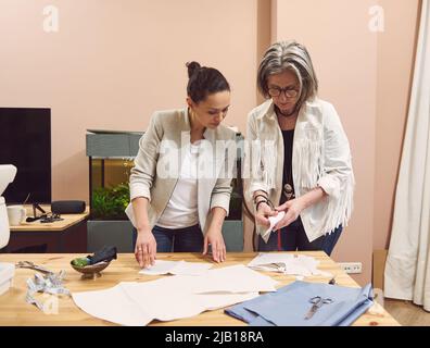 Two pretty seamstresses of different nationalities and ages in a tailoring workshop discussing fabric, sewing pattern and sketch during production pro Stock Photo