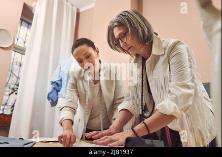 Inspired and skilled team of two diverse women fashion designers seamstresses working with sewing pattern , creating new collection of clothes in a cl Stock Photo