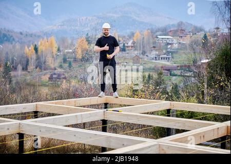 Man worker building wooden frame house on pile foundation. Portrait of carpenter measuring wooden planks and showing thumbs up. Carpentry concept. Stock Photo