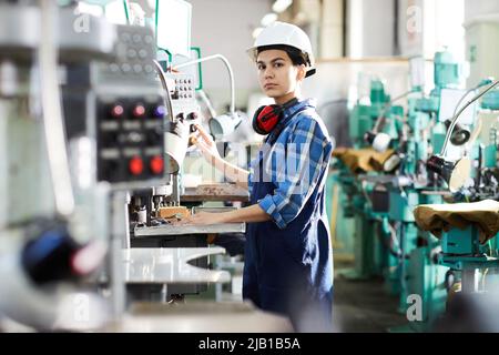 Serious thoughtful woman engineer in uniform standing at industrial machine and producing small gear for wristwatch, she working at production line Stock Photo