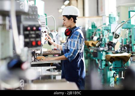 Serious young female engineer in hardhat concentrated on production process pushing button on control panel of industrial machine Stock Photo