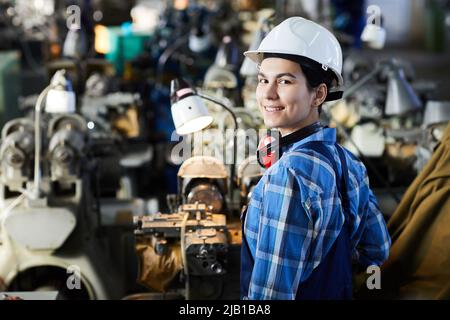 Cheerful confident attractive woman in construction hardhat standing at lathe machine and turning back while looking at camera, woman in industry conc Stock Photo