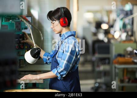 Serious skilled woman machinist in ear protectors and safety goggles pushing button while operating machine in workshop Stock Photo