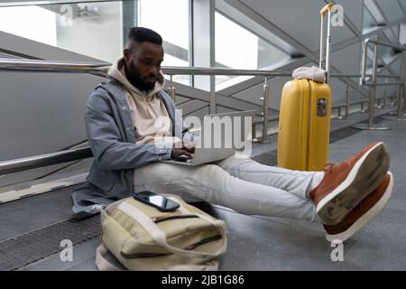 Intent young black guy with computer on laps sitting on airports floor booking apartment for journey Stock Photo