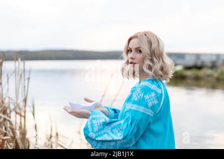 Young beautiful Ukrainian woman in a blue embroidered shirt, holding a paper boat made of white paper and looking forward with hope. Stock Photo